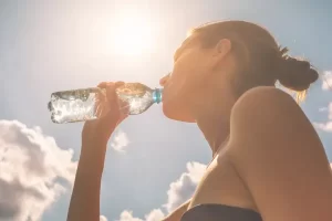 Mulher bebendo água sob a luz do sol, destacando a importância da hidratação para o corpo.